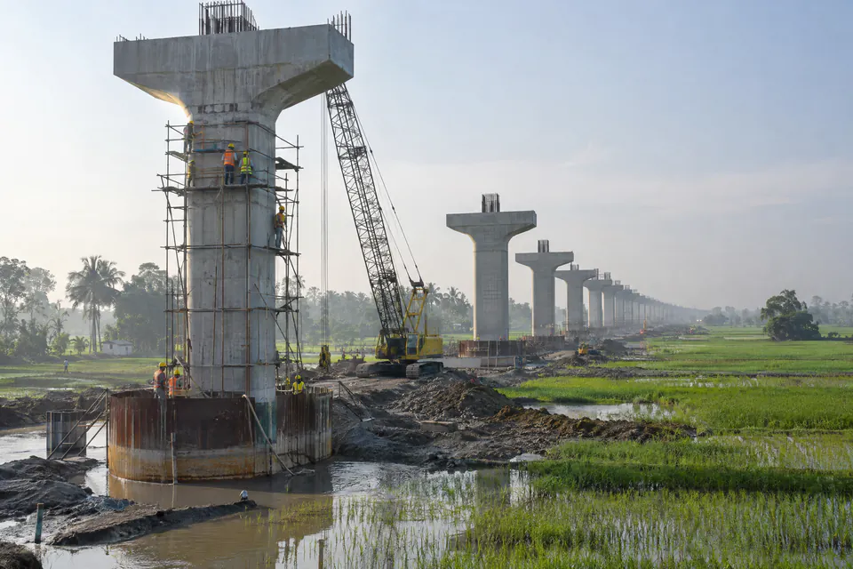Illustration: an elevated rail viaduct under construction across rice fields, with steel scaffolding at a pier