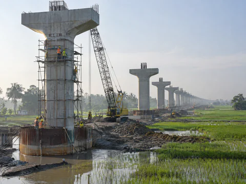 Illustration: an elevated rail viaduct under construction across rice fields, with steel scaffolding at a pier