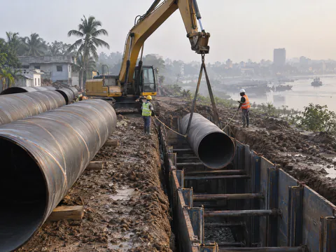 Illustration: large spiral welded steel pipes being lowered into a shored trench for a city water main in Bangladesh